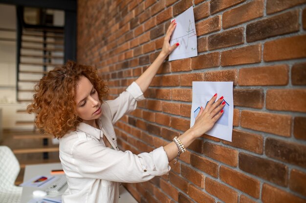 Zdjęcie imbirowa kobieta umieszcza diagramy na ścianie w biurze