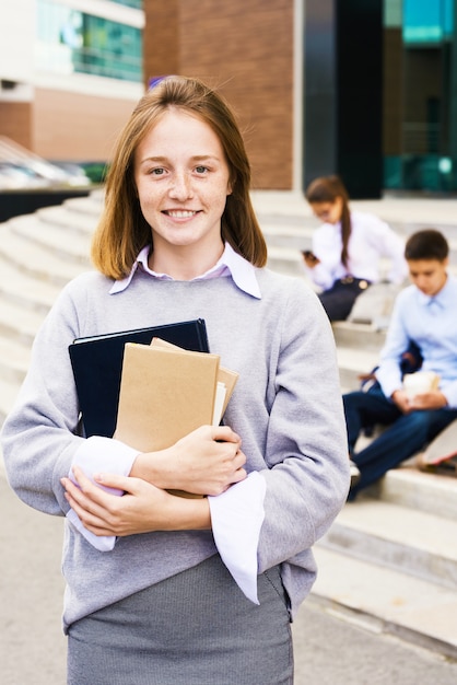 Imbirowa uczennica uśmiecha się szczęśliwie stojąc z podręcznikami przed budynkiem szkoły