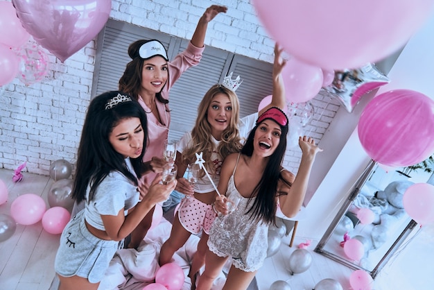 Impreza dla pań! Widok z góry czterech atrakcyjnych młodych uśmiechniętych kobiet w piżamach pijących szampana podczas piżama party w sypialni