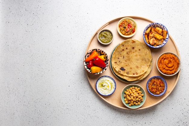 Indian Thali - wybór różnych dań podawanych na okrągłym drewnianym półmisku, biały stół. Różne indyjskie wegetariańskie meze z chlebem chapati, paneer, dal, chutney w miseczkach. Widok z góry, miejsce na tekst