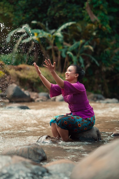 Indonezyjska kobieta siedzi na małym kamieniu i bawi się wodą, podczas gdy jej sukienka zamoczyła się w rzece