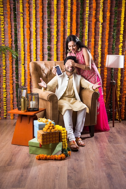 Indyjska atrakcyjna para w tradycyjnym stroju świętującym festiwal Diwali, urodziny lub rocznicę z prezentami niespodziankami i słodkim laddoo na tle zdobionym kwiatami nagietka