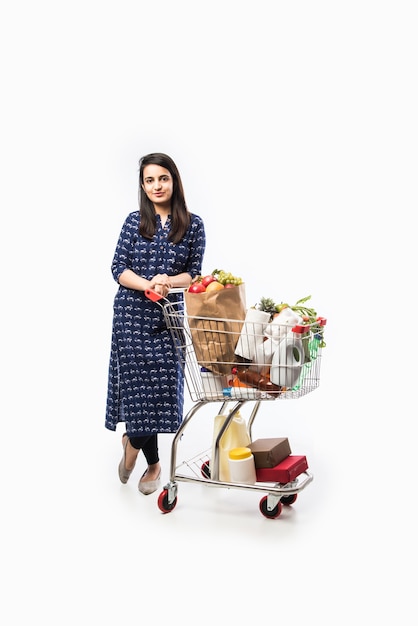 Indyjska młoda kobieta z wózkiem na zakupy lub wózkiem pełnym artykułów spożywczych, warzyw i owoców. Izolowane Zdjęcie pełnej długości na białej ścianie