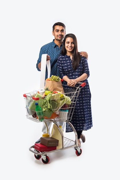 Indyjska młoda para z wózkiem na zakupy lub wózkiem pełnym artykułów spożywczych, warzyw i owoców. Izolowane Zdjęcie pełnej długości na białej ścianie