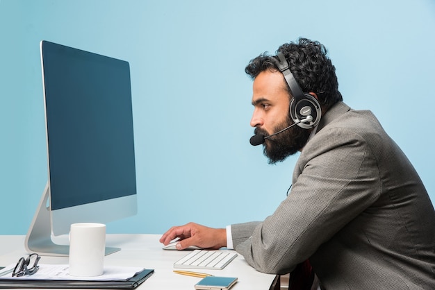 Indyjski azjatycki brodaty młody mężczyzna w garniturze w call center, w pomieszczeniu, słuchając słuchawek, przeglądając komputer lub prowadząc rozmowę głosową