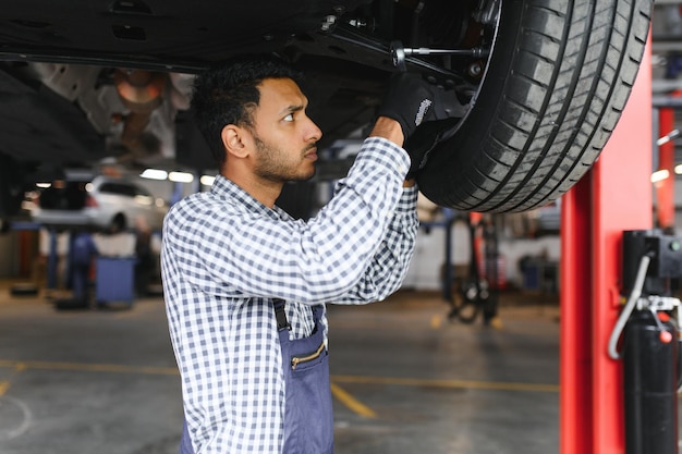 Indyjski mechanik samochodowy stojący i pracujący na stacji paliw Specjaliści samochodowi badający podniesiony samochód Profesjonalni mechanicy w mundurze mechanika w kolorze niebieskim