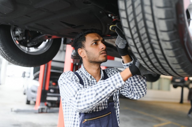 Indyjski mechanik samochodowy stojący i pracujący na stacji paliw Specjaliści samochodowi badający podniesiony samochód Profesjonalni mechanicy w mundurze mechanika w kolorze niebieskim