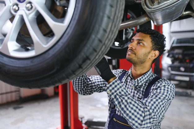 Indyjski mechanik samochodowy stojący i pracujący na stacji paliw Specjaliści samochodowi badający podniesiony samochód Profesjonalni mechanicy w mundurze mechanika w kolorze niebieskim