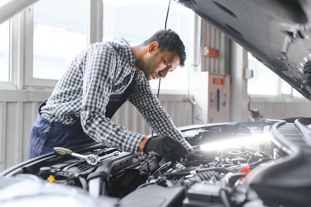 Indyjski mechanik samochodowy stojący i pracujący na stacji paliw Specjaliści samochodowi badający podniesiony samochód Profesjonalni mechanicy w mundurze mechanika w kolorze niebieskim