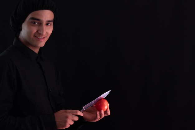 Indyjski mężczyzna kucharz pozuje uśmiechając się do kamery na czarnym tle ubrany w mundur i trzymający w dłoni nóż, którym kroi warzywo