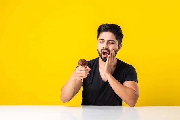 Indyjski młody mężczyzna jedzący lody w rożku i mający ból zębów z powodu ubytku, siedzący przy stole na żółtym lub białym tle