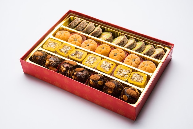 Indyjskie słodycze lub mithai w pudełku i lampach oliwnych lub diya z kwiatami na ozdobnym lub kolorowym tle, selektywna ostrość