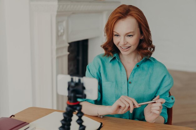 Influencer w mediach społecznościowych, bloger, który trzyma telefon komórkowy na statywie, z radością patrzy na strumienie z kamery, kręci podcasty online, vlog siedzi przy komputerze, prowadzi komunikację online lub czat używa specjalnej aplikacji