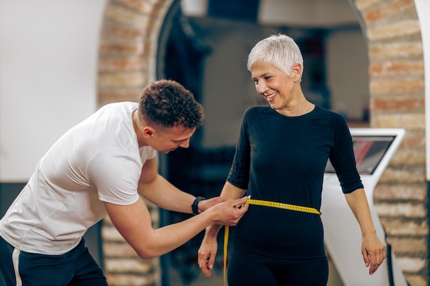 Instruktor fitness pomiaru talii starszej kobiety przed treningiem na siłowni.