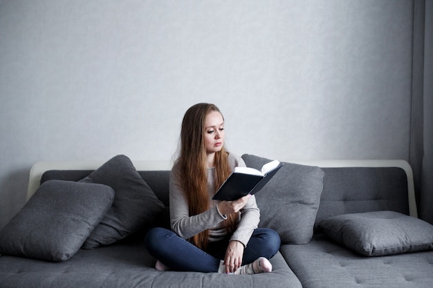 Inteligentne młoda kobieta siedzi na kanapie w domu, czytając książkę