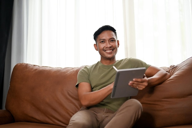 Inteligentny azjatycki mężczyzna używający cyfrowego tabletu podczas relaksu na sofie w salonie