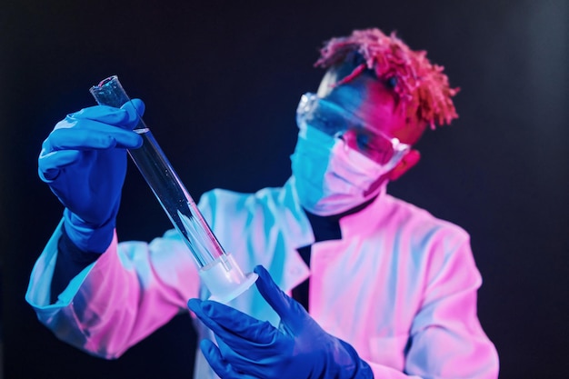 Inteligentny naukowiec w ochronnym mundurze trzymającym probówkę Koncepcja koronawirusa Futurystyczne oświetlenie neonowe Młody mężczyzna z Afryki w studio