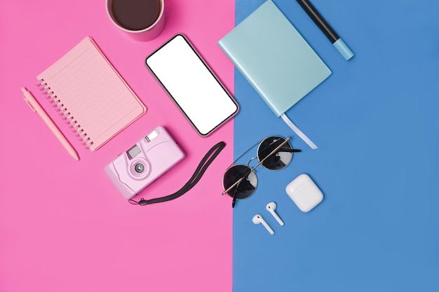 Inteligentny telefon, aparat fotograficzny, notebook, bezprzewodowe słuchawki i okulary przeciwsłoneczne na niebieskim i różowym tle.