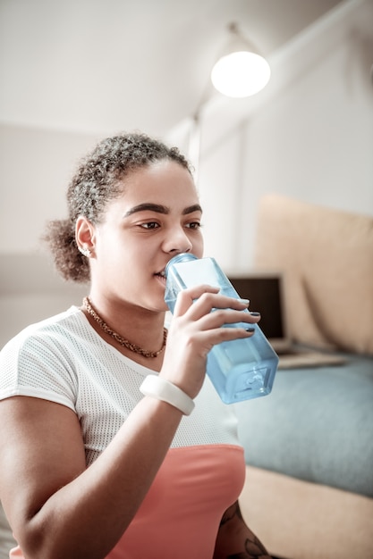 Intensywny trening sportowy. African American miło kobieta pije czystą wodę z niebieskiej plastikowej butelki