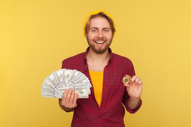 Inwestycja cyfrowa, wymiana pieniędzy. Wesoły hipster facet w kraciastej koszuli, trzymając banknoty dolarowe i bitcoin, kryptowaluta monety btc, uśmiechając się do kamery. studio strzał na białym tle na żółtym tle