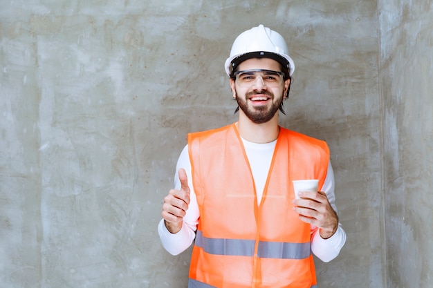 Inżynier człowiek w białym kasku i okulary ochronne, trzymając kubek napoju i pokazując znak przyjemności.