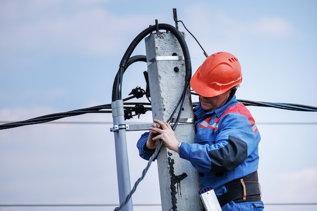 Inżynier Elektryk Wykonuje Okablowanie Na Wysokim Słupie Stojącym Na Schodach. Prace Elektryczne Na Wysokościach. Dom Podkluchenie Do Linii Energetycznych