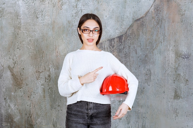 Inżynier kobieta trzyma czerwony hełm i pokazuje coś dookoła.