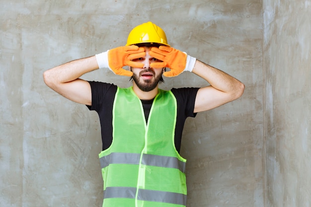 Inżynier w żółtym kasku i przemysłowych rękawiczkach patrzący przez palce