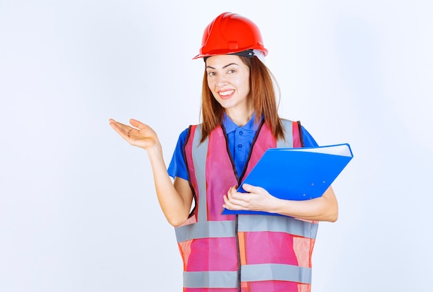 Inżynierka w czerwonym hełmie trzymająca niebieski folder projektu i przedstawiająca kogoś lub coś.