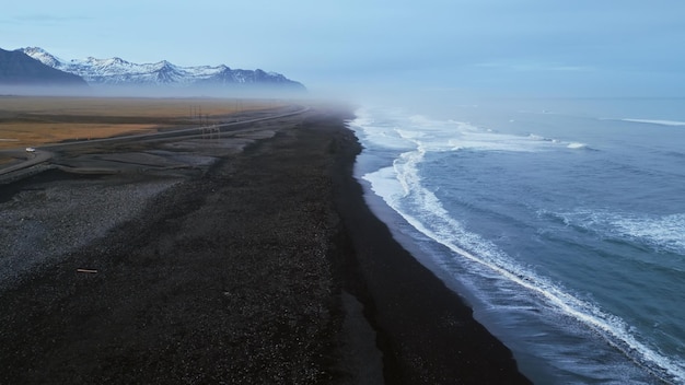 Islandzka czarna piaszczysta plaża nakręcona dronem, piękna linia brzegowa z falami rozbijającymi się na wybrzeżu Atlantyku. Skandynawski krajobraz z śnieżnymi górami i czarnym piaskiem na plażach, malownicza trasa.