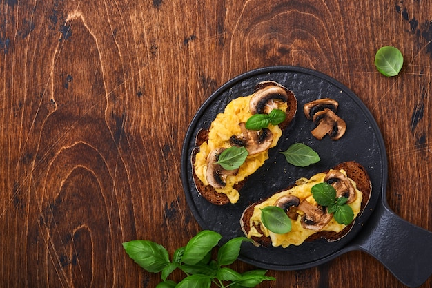 Jajecznica Ze Smażonymi Pieczarkami I Bazylią Na Chlebie Na Tle Czarnego Stołu. Domowe śniadanie Lub Brunch - Jajecznica I Kanapki Z Grzybami. Widok Z Góry Z Miejscem Na Kopię