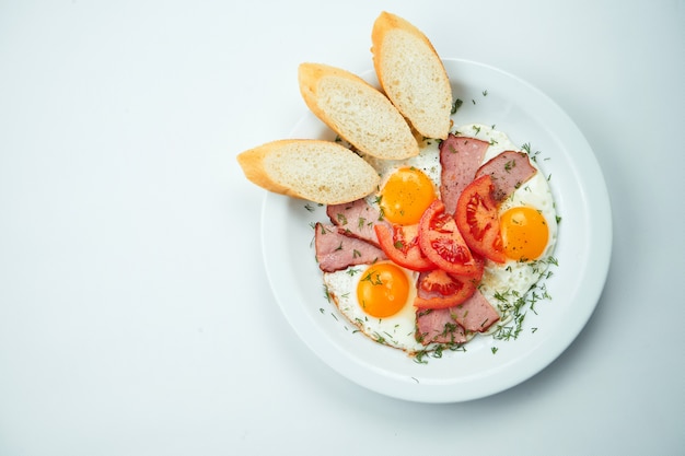 Jajka sadzone z pomidorami, chlebem i szynką na białym talerzu na szarym stole. Widok z góry z miejsca kopiowania