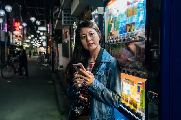 Japonka, opierając się na automatach w Japonii przy użyciu telefonu komórkowego, relaks. Młody student lub turystka z aparatem wysyłanie SMS-ów przez telefon stojący obok sprzedawcy samochodów. dziewczyna na zewnątrz w nocy osaka jp