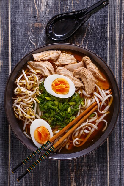 Japońska zupa ramen z kurczakiem, jajkiem, szczypiorkiem i kiełkami na ciemnym tle drewnianych.