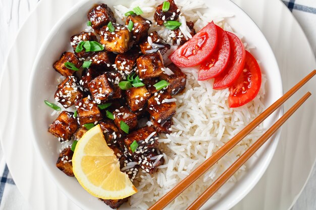 Japońskie teriyaki pieczone tofu z ryżem posypane szczypiorkiem, sezam podawane z ryżem, pomidor pokrojony w plastry, cytryna na białej misce z pałeczkami na białej drewnianej powierzchni, widok z góry, układanie na płasko