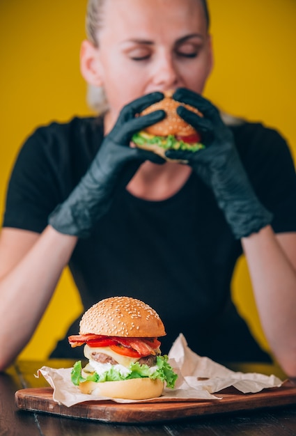 Jasne soczyste apetyczne burgery z kotletem, serem, marynowanymi ogórkami, pomidorami i boczkiem w rękach dziewczyny, dziewczyny w specjalnych rękawiczkach do burgerów