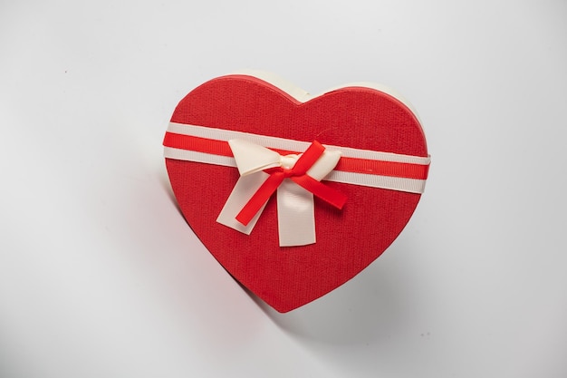 Jasnoczerwone satynowe pudełko w kształcie serca przewiązane białą wstążką z kokardką na białym tle
