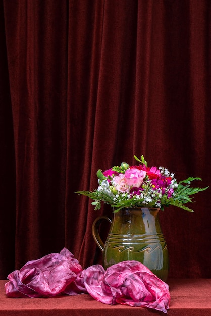 Jasny bukiet kwiatów w zielonym wazonie na tle ciemnej tkaniny obok różowego szalika