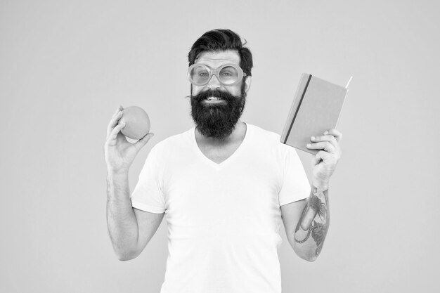 Jasny i żywy Mężczyzna brodaty wesoły uśmiechnięty hipster nosi pomarańczowe okulary przeciwsłoneczne, trzymając pomarańczowe owoce i książkę Notatnik Całkowita pomarańcza Facet z długą brodą lubi kolor pomarańczowy Źródło witamin