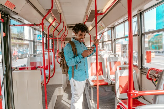 Jazda autobusem. Wesoły ciemnoskóry facet patrzący na smartfona trzymającego poręcz stojący w pustym autobusie miasta