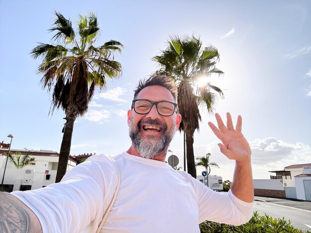 Jeden dojrzały mężczyzna robi selfie na świeżym powietrzu, spacerując po ulicy w tropikalnym, malowniczym miejscu z palmami na tle. Szczęśliwy turysta komunikuje się za pomocą komórkowego telefonu komórkowego.