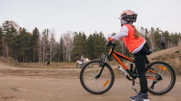 Jeden kaukaski dzieci jeździ po torze rowerowym w brudnym parku Dziewczyna jedzie na czarno pomarańczowy cykl na torze wyścigowym Dzieciak idzie uprawiać sporty rowerowe Jazda ruchowa na rowerze z plecakiem i kaskiem Twardy ogon roweru górskiego