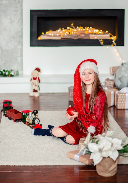 Jeden ładny wesoły dziewczyna santa hat w pobliżu choinki. Trzyma czerwoną piłkę Bożego Narodzenia.