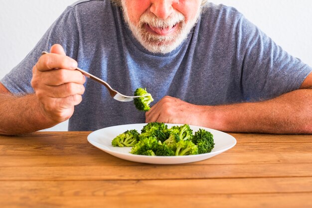 Jeden starszy patrzący z obrzydzeniem na brokuły na talerzu na drewnianym stole w domu - próbuje zrobić dietę i schudnąć