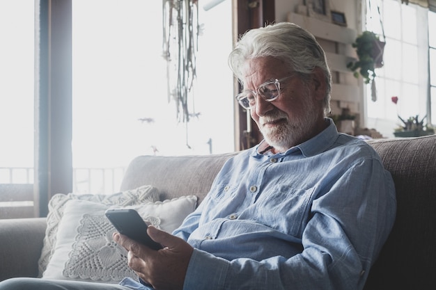 Jeden Stary I Dojrzały Mężczyzna Za Pomocą Telefonu, Praca Z Smartphone, Siedząc Na Kanapie W Domu Kryty Koncepcja I Styl życia Człowieka Biznesu. Mężczyzna Emeryt I Emeryt Bawią Się Relaksując Się Na Kanapie