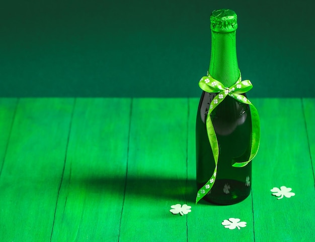 Jedna butelka zielonego piwa z wstążką związaną w łuku i trzy papierowe koniczyny na drewnianym tle
