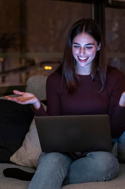 Jedna kobieta uśmiecha się i bawi się podczas rozmowy wideo za pomocą laptopa siedząc wygodnie w domu na kanapie w ciemny wieczór ludzie rozmawiają na notebooku z połączeniem internetowym