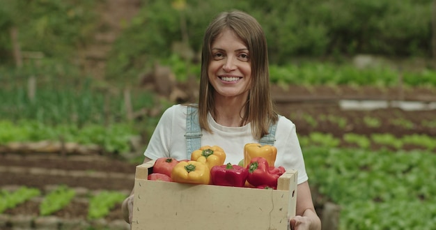 Jedna szczęśliwa młoda rolniczka trzymająca kosz organicznych warzyw stojąca w lokalnej farmie Zbliżenie twarzy kobiety-rolnika trzyma w ręku żółtą i czerwoną paprykę