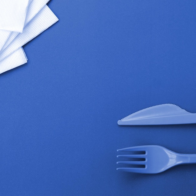 Jednorazowe plastikowe sztućce Plastikowy widelec i nóż leżą na fantomowej klasycznej niebieskiej powierzchni tła obok serwetek