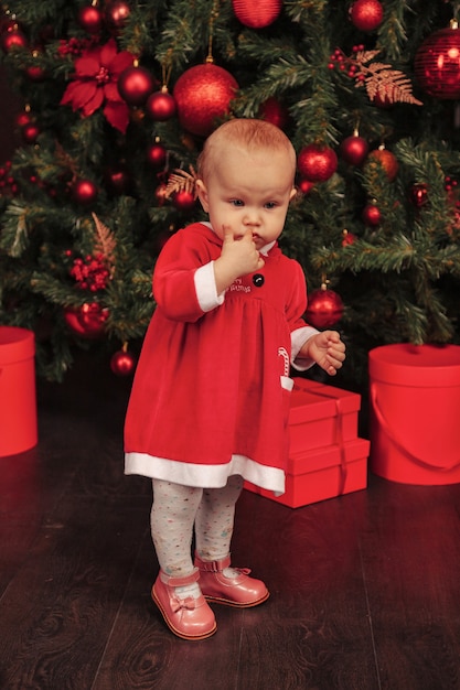 Jednoroczne dziecko o niebieskich oczach w stroju świątecznym bawiące się w salonie z drzewami i pudełkami na prezenty. Słodkie dziecko z emocjami wieczoru wakacyjnego. Koncepcja rodzinnych obchodów Świąt Bożego Narodzenia i Szczęśliwego Nowego Roku